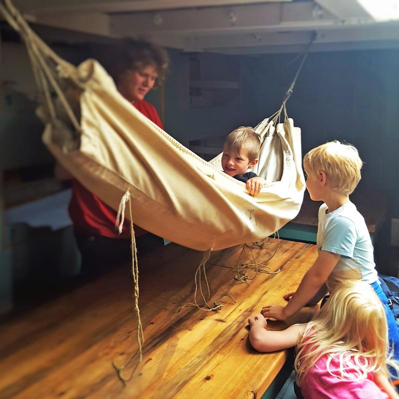 Hengekøye ombord skip, ett barn i hengekøya, to barn utenfor, én voksen bak. (Foto/Photo)