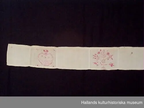 Smalt hängkläde ihopsytt av tygbitar av vitt linne, där varannan tygbit är broderad med hallandssömsmönster i rött (rosa) bomullsgarn. Sju partier med fyra olika mönster, samt initialer SLB.