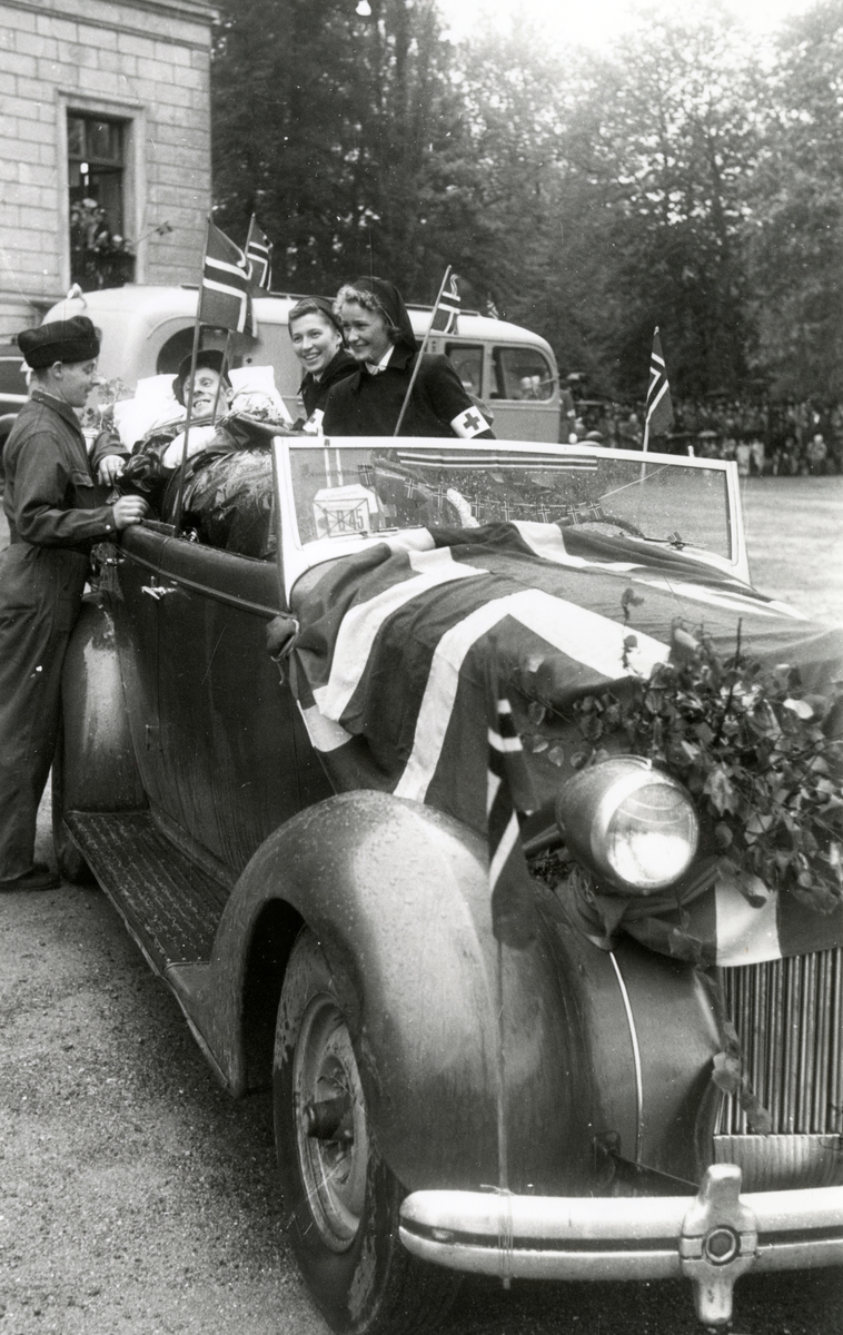 Fra fredsdagene i 1945, antagelig 17.mai.
På Slottsplassen med en såret hjemmefrontkar liggende i bil, av typen Packard 1939, på båre med blomster i hendene. En uniformert mann og  to kvinnelige sykelpeiere ved bilen, som er pyntet med løv og norske flagg.