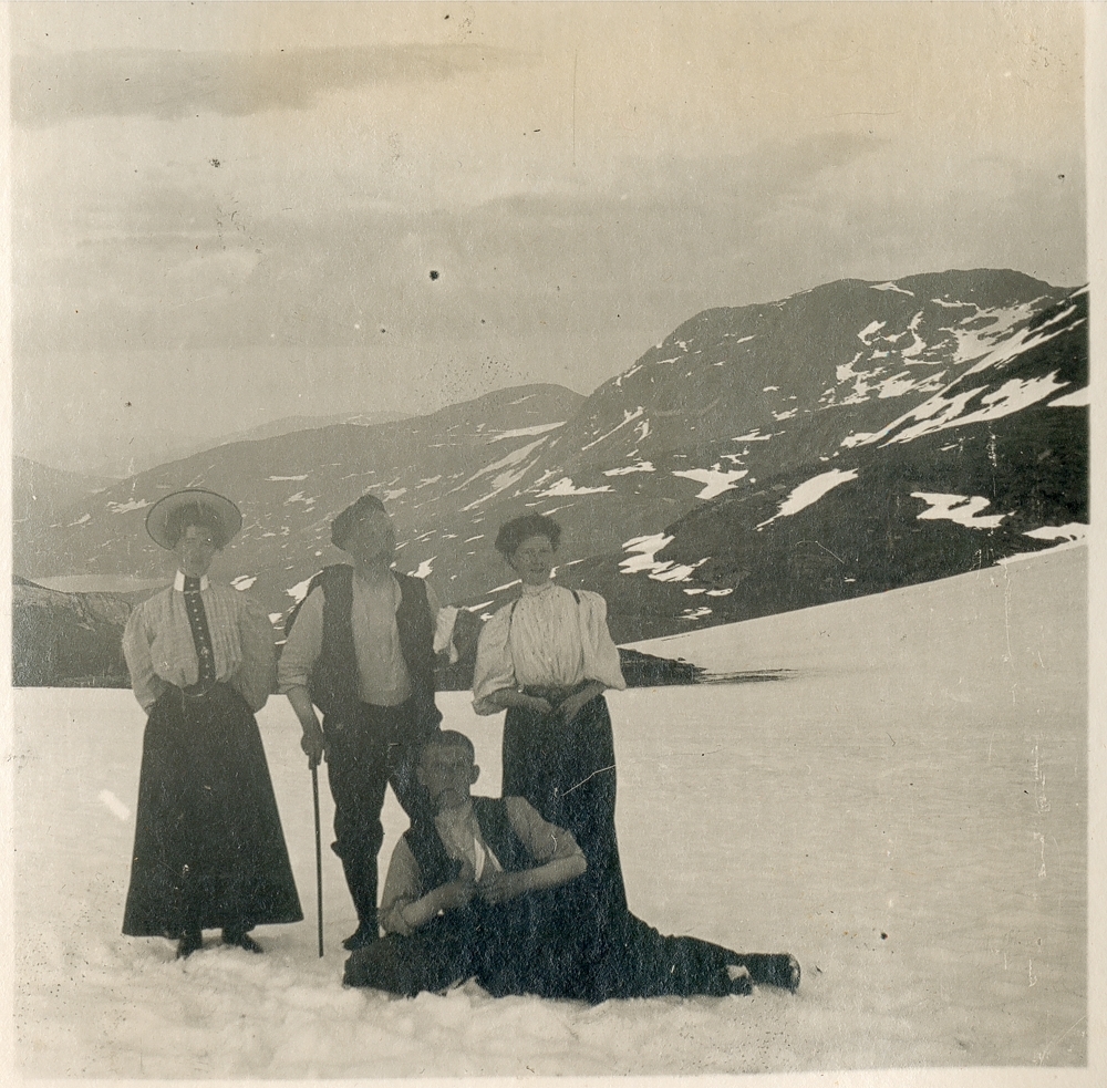 Gruppe, ute. Snø. Fjelltur. Utsiktsbilde. To kvinner med skjørt og to menn. Den ene mannen ligger foran i snøen.