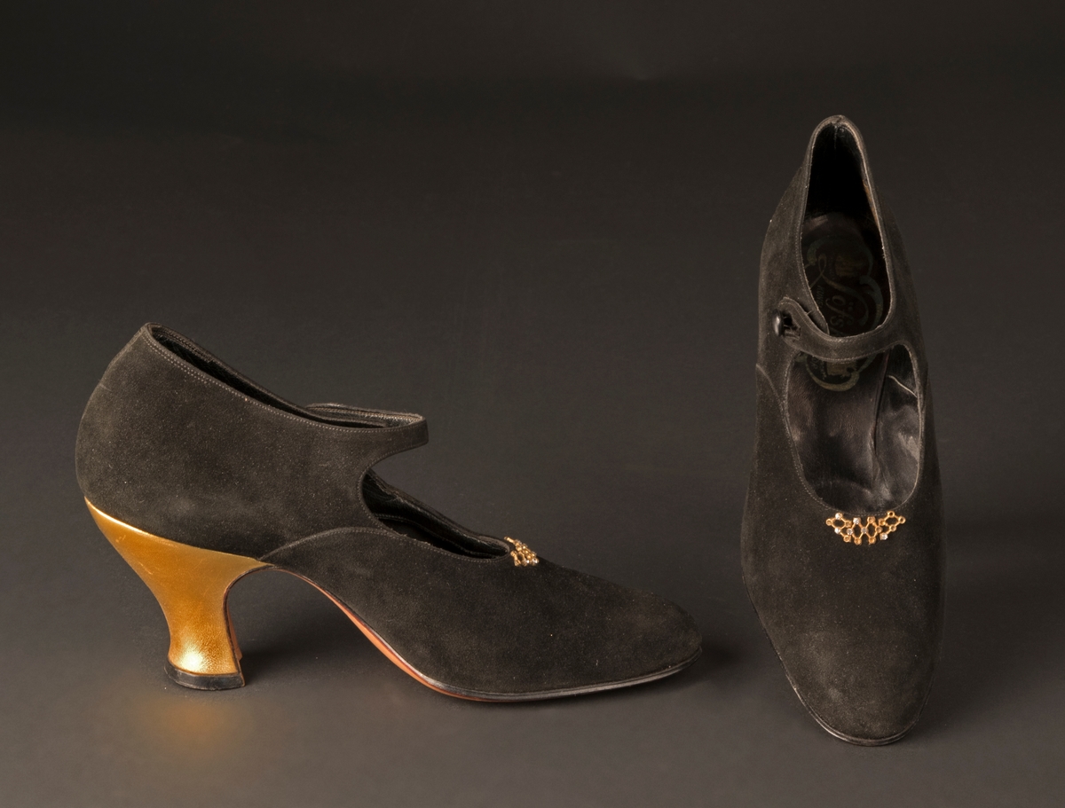 Ett par damskor från mitten av 1920-talet av svart mocka, klacken klädd med guldchevreau. Vristrem, som knäpps i sidan. Gulmetallspänne med vit strass monterad mitt på skon. Helfodrad. Lädersula. Storlek 36.
