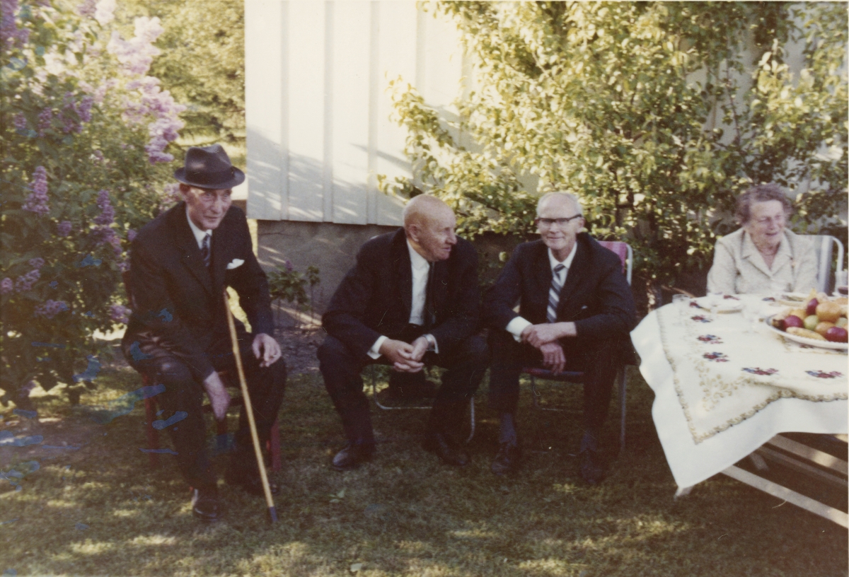 Tre eldre menn med dress og en dame sitter i hagen på Braate gård.