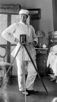 Fotograf i hvit dress og med hvit lue poserer bak et gammelt kamera på stativ.