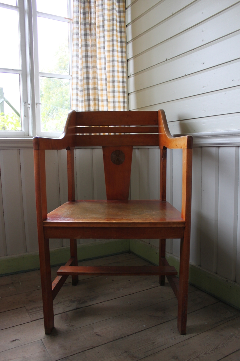 Karmstol i trä med tillhörande sittdyna. Stolen är försedd med armstöd och har snidade trädekorer på ryggstöd och ryggstödets sidor.
