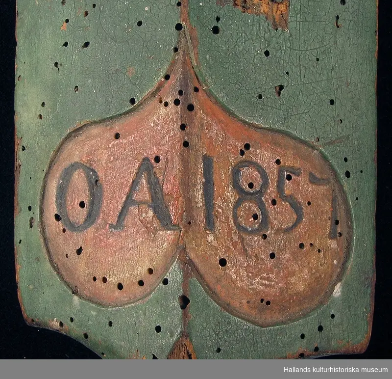 Klappträ som saknar handtag. Föremålet är dekorerat med utskärningar i form av en karvsnittsrosett. Målat i grön bottenfärg med detaljer i rött och gråvitt. Märkt med årtal och monogram 1857 OA, inom en hjärtform.