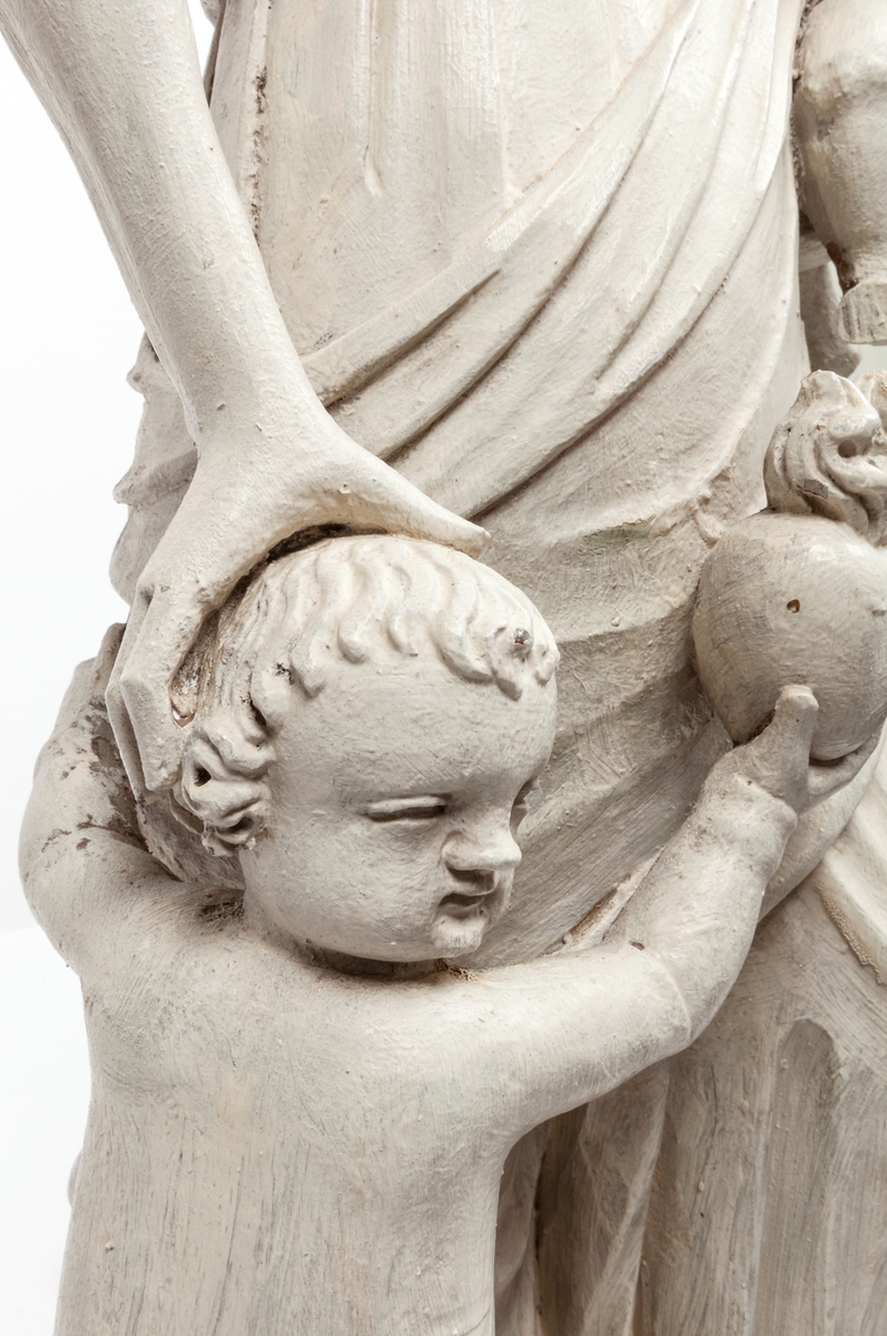 Kyrklig skulptur, trä, staty från gamla korskranket i Heliga Trefaldighets kyrka i Gävle, av Ewert Friis omkring 1665. 

Stående kvinnofigur med naket barn på vänster arm samt stående naket barn vid höger sida. Sistnämnda barn har ett brinnande hjärta i höger hand och kvinnans vänstra hand på sitt huvud. Vänster ben är främst, foten avslagen. Sittande barnets ena ben och andra fot avslagna. 
Fyrsidigt, profilerat postament. 
Gråvit färg.
