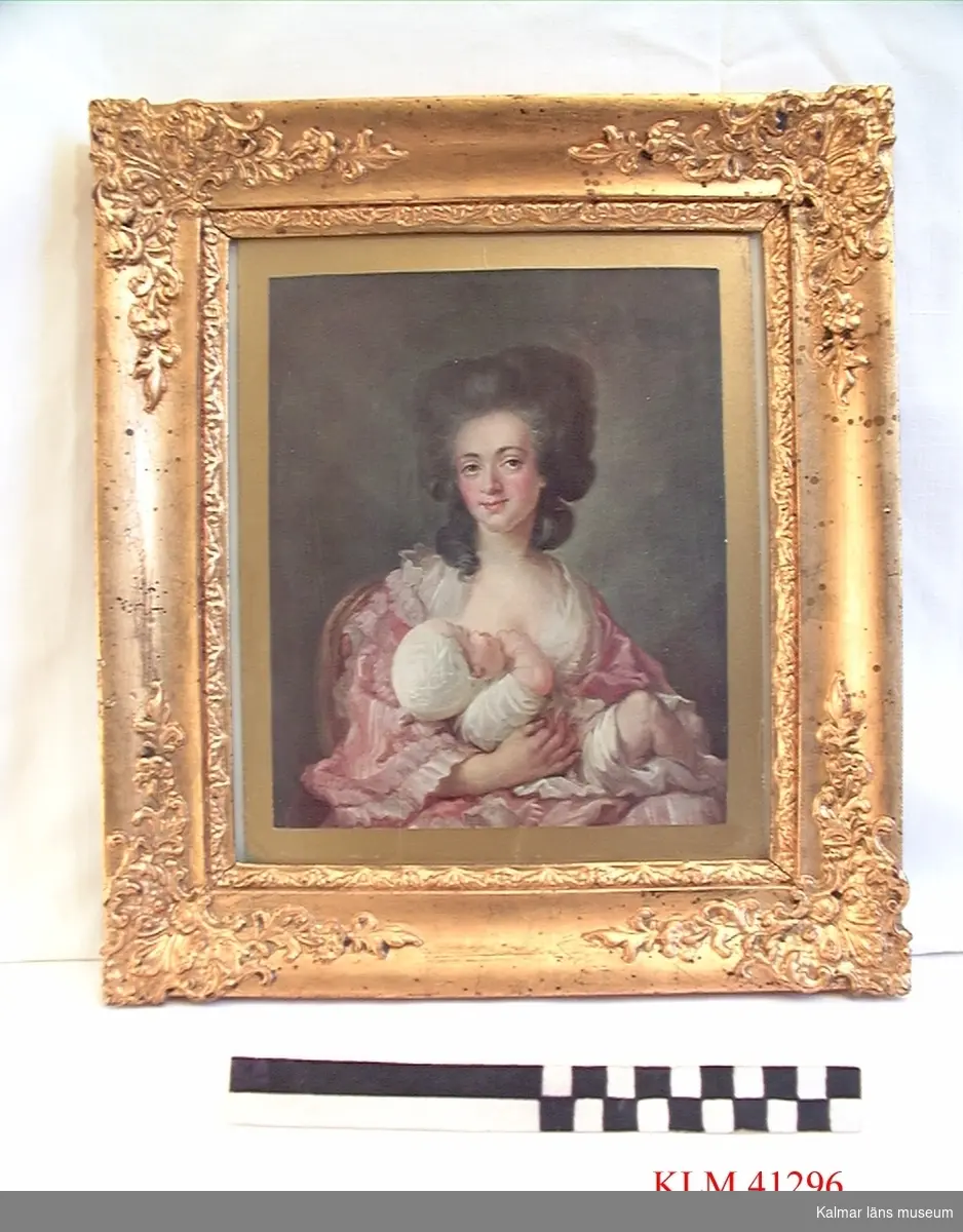 KLM 41296 Tavla, papper. Tryck på papper i bred ram av förgyllt trä, stående format. Bröstbild av kvinna klädd i rosa klänning av rokokosnitt med spädbarn i famnen.