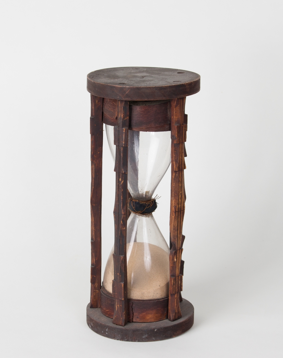 Timeglass består av to stk. glassbeholder med en smal kanal imellom som fin sand renner igjennom. Står i stativ laget av tre.
