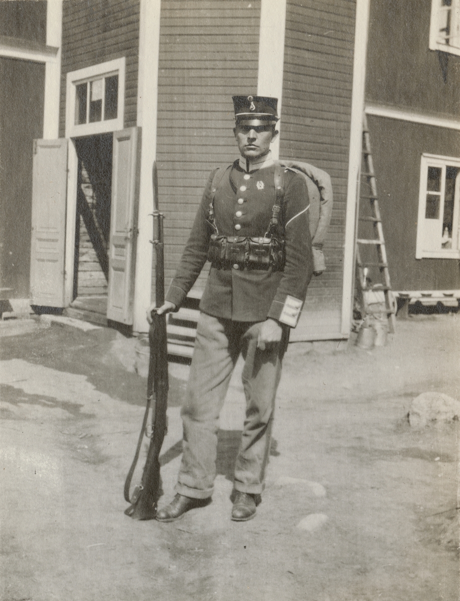 En soldat från Göta livgarde I 2 med gevär och ryggsäck framför ett trähus.