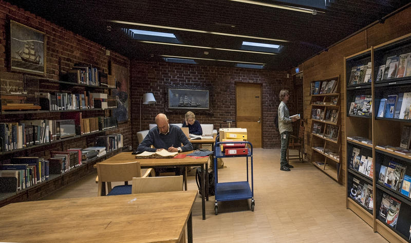 To personer sitter på lesesalen og en person står ved bokhylle på biblioteket. (Foto/Photo)