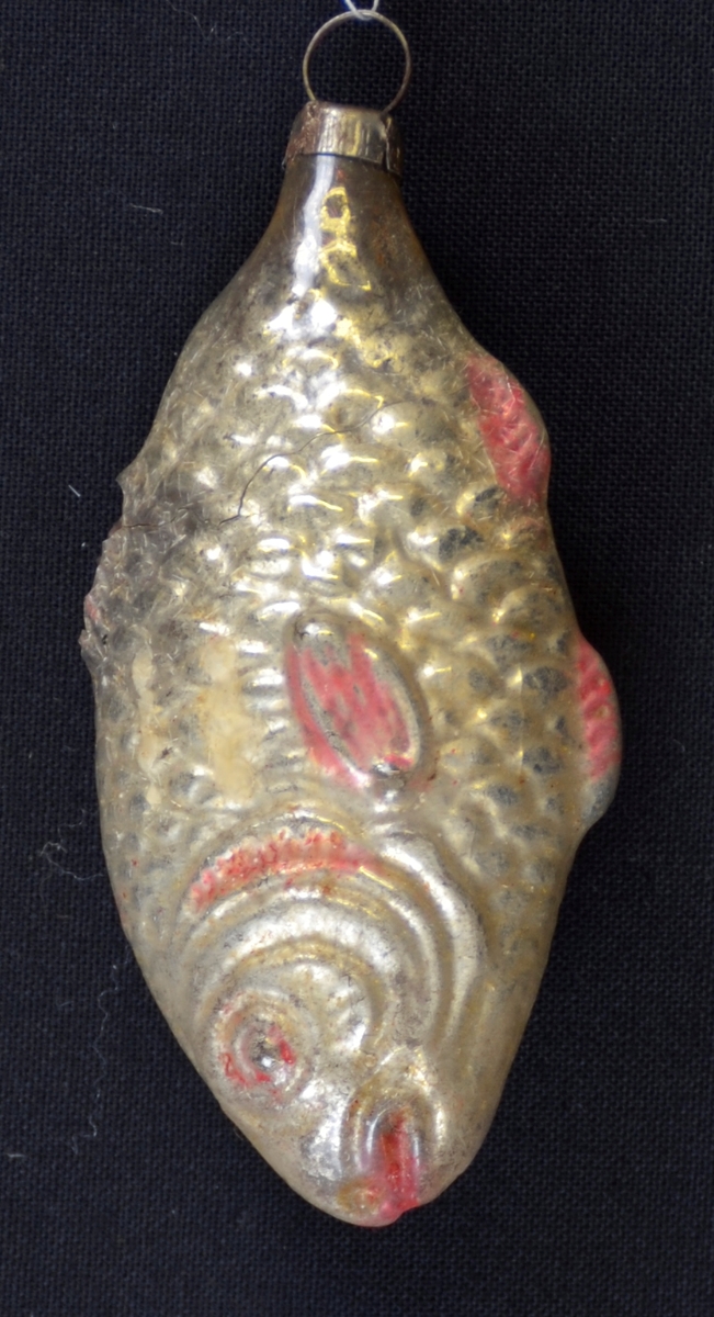 Juletrepynt i glas. Hol. Forma er ein relativt naturalistisk fisk. Sølv /gull med noko raudt. Metalloppheng med ring festa i stjerten.