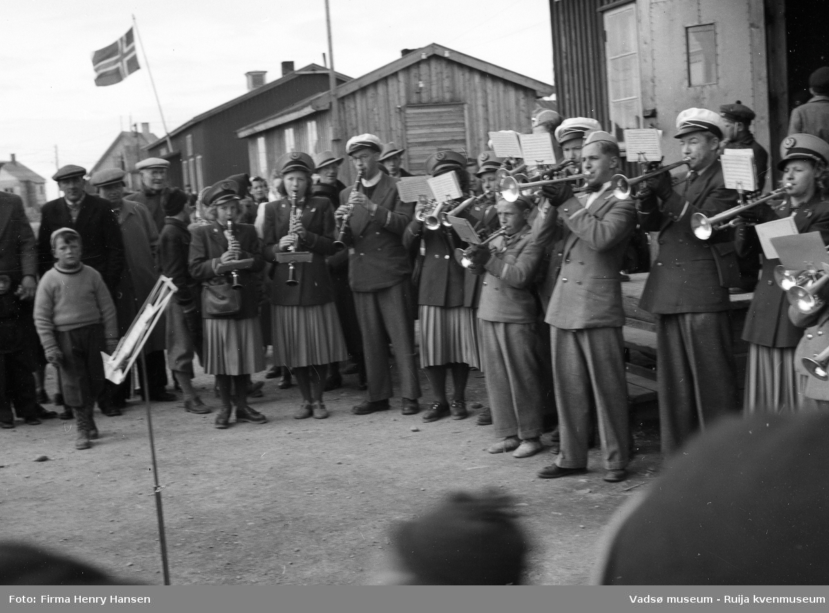 Vadsø sentrum 17. mai 1951, Musikkorps står oppstilt og spiller foran postbrakka. Voksne og barn er tilskuere. Vi ser deler av fire andre bygninger, to bolighus og en brakke.