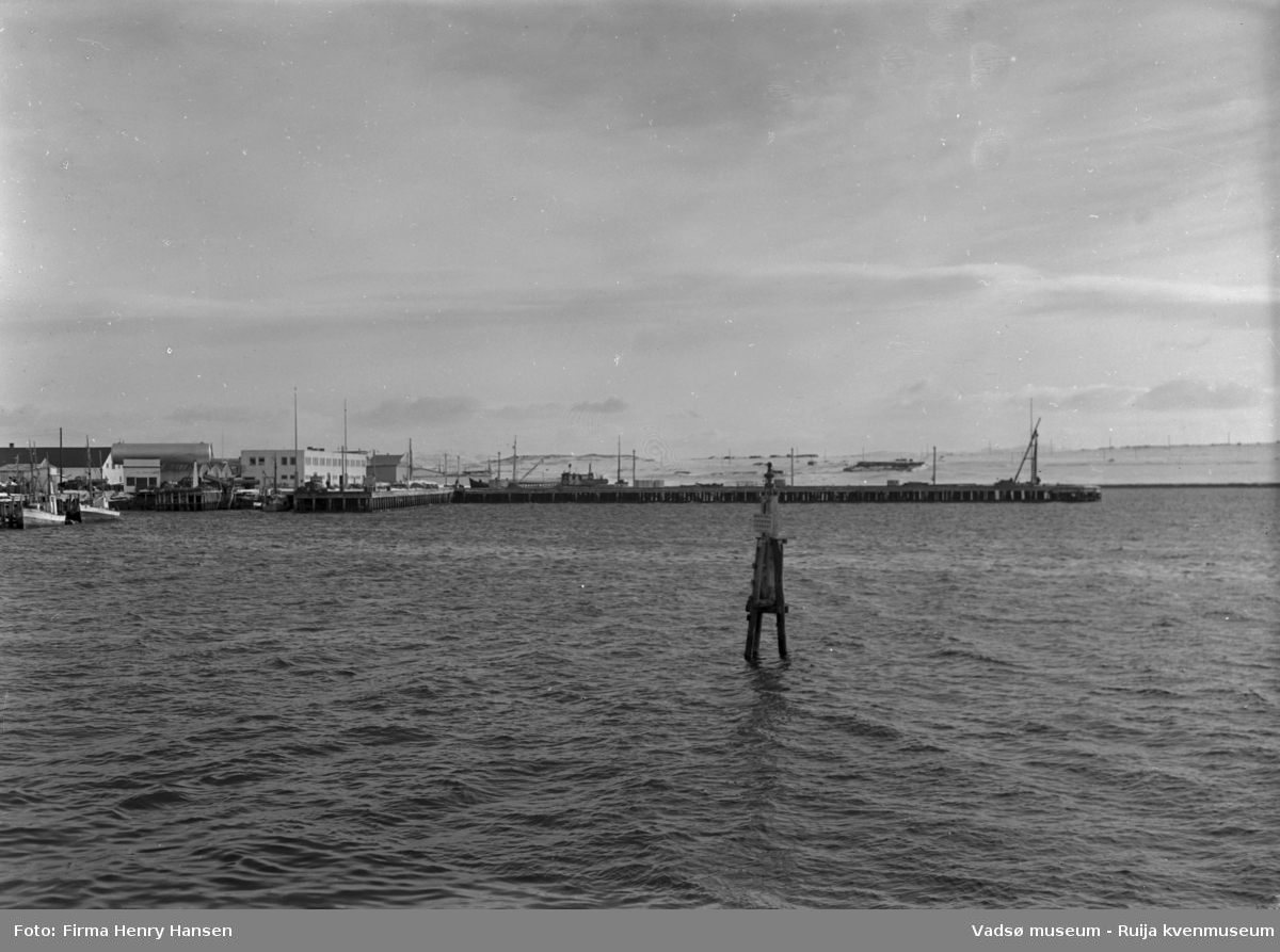 Vadsø april 1952. Bildet er tatt fra fastlandet mot syd. Vi ser havna med Dampskipskaia og dampskipsekspedisjonen midt i bildet. Videre ser vi flere kaianlegg, fiskebåter, tanker, bygninger. I bakre billedkant ser vi Vadsøya og noen bygninger.