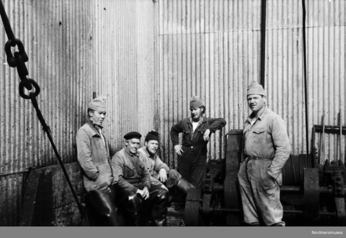 Foto fra Grytviken i Sør-Georgia, og fra en norsk hvalstasjon der, og viser en gruppe på fem menn i arbeidsklær. Bildet kan dateres mellom 1959 til 1960. Fotograf er trolig Rolf Karlsen. Pause ved en av de store vinsjene. Fra Nordmøre museums fotosamlinger.