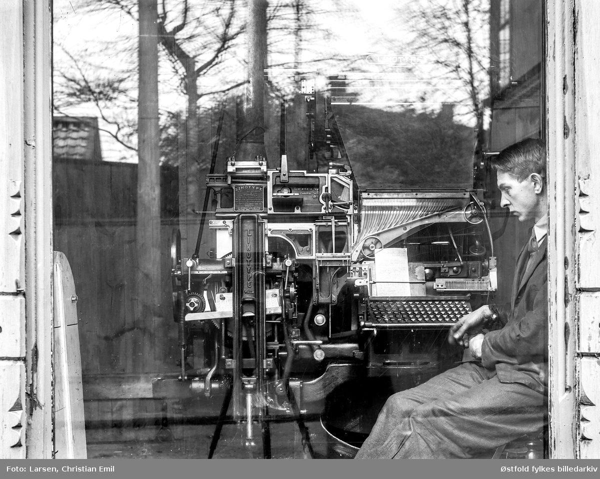 Avisa Sarpens trykkeri i Sarpsborg, mann passer trykkerimaskin, fotografert gjennom vindusrute.