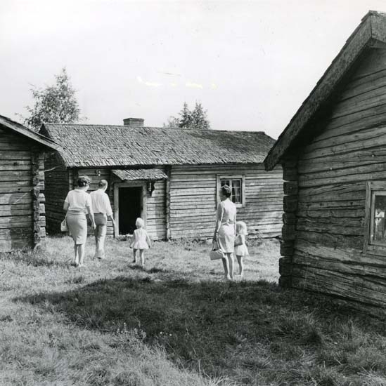Övre Älmesbovallen, Edsbyn 1970. Vuxna och barn går i gräset mellan fäbodstugor.