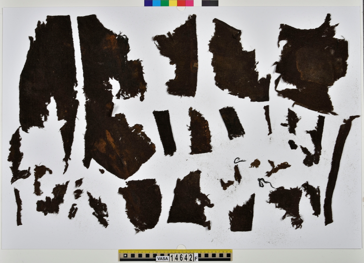 Textil.
132 fragment uppdelade på fyndnummer 14642a-f.
Fnr 14642a består av 42 fragment av ull vävda i tuskaft. Tyget har varit valkat på ena sidan. Många av fragmenten har bevarade originalkanter med sömmar och fållar.
Fnr 14642b består av 2 fragment av ull vävt i 2/1-kypert. Fragmentet har en bevarad originalkant.
Fnr 14642c består av 14 fragment av ull vävda i 2/2-kypert. Flera av fragmenten har en bevarad originalkant.
Fnr 14642d består av 28 fragment av ull vävda i 2/1-kypert. Fyra av fragmenten har en bevarad originalkant. Flera av fragmenten består bara av lösa trådar.
Fnr 14642e består av 9 fragment (4 större samt 5 mycket små) av ull vävda i tuskaft. Tyget har varit valkat på ena sidan. Fragmenten har bevarade originalkanter med spår av sömmar.
Fnr 14642f består av 37 fragment av ull vävda i tuskaft. Flera av fragmenten har bevarade originalkanter med fållar och spår av sömmar.