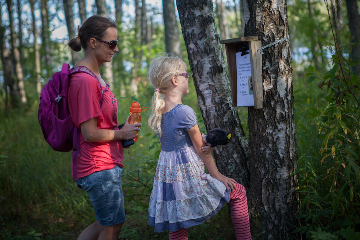 Mor og datter leser på en naturstipost på Blikomøya. Mor har olabukse og t-skjorte på seg og datteren har sommerkjole (Foto/Photo)