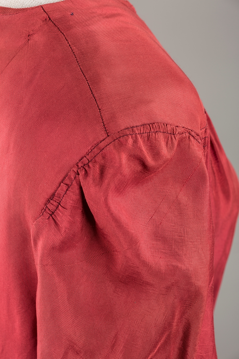 Form: Glatt, klokkeformet skjørt. Forstykkene rynket til buede bærestykker foran. Overdel rynket til skjørt. Korte ermer. Lukket med trykknapper. Belte fastsydd.