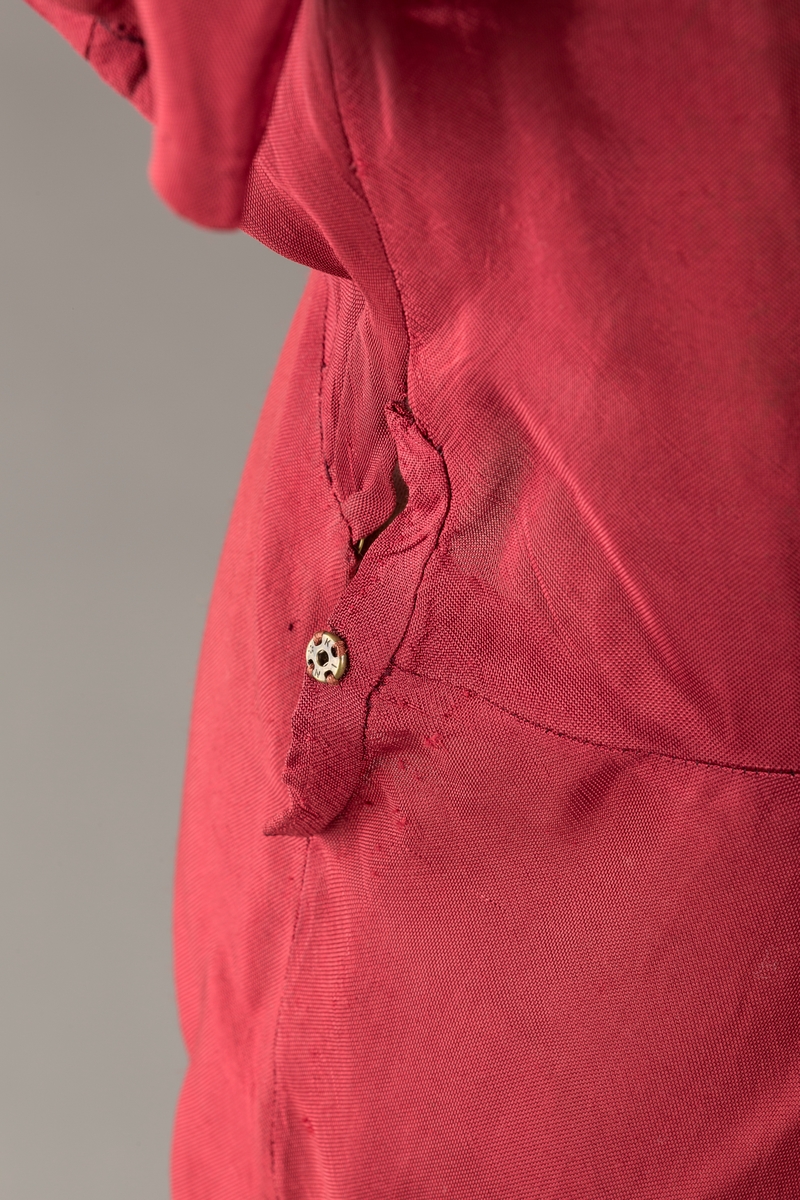 Form: Glatt, klokkeformet skjørt. Forstykkene rynket til buede bærestykker foran. Overdel rynket til skjørt. Korte ermer. Lukket med trykknapper. Belte fastsydd.