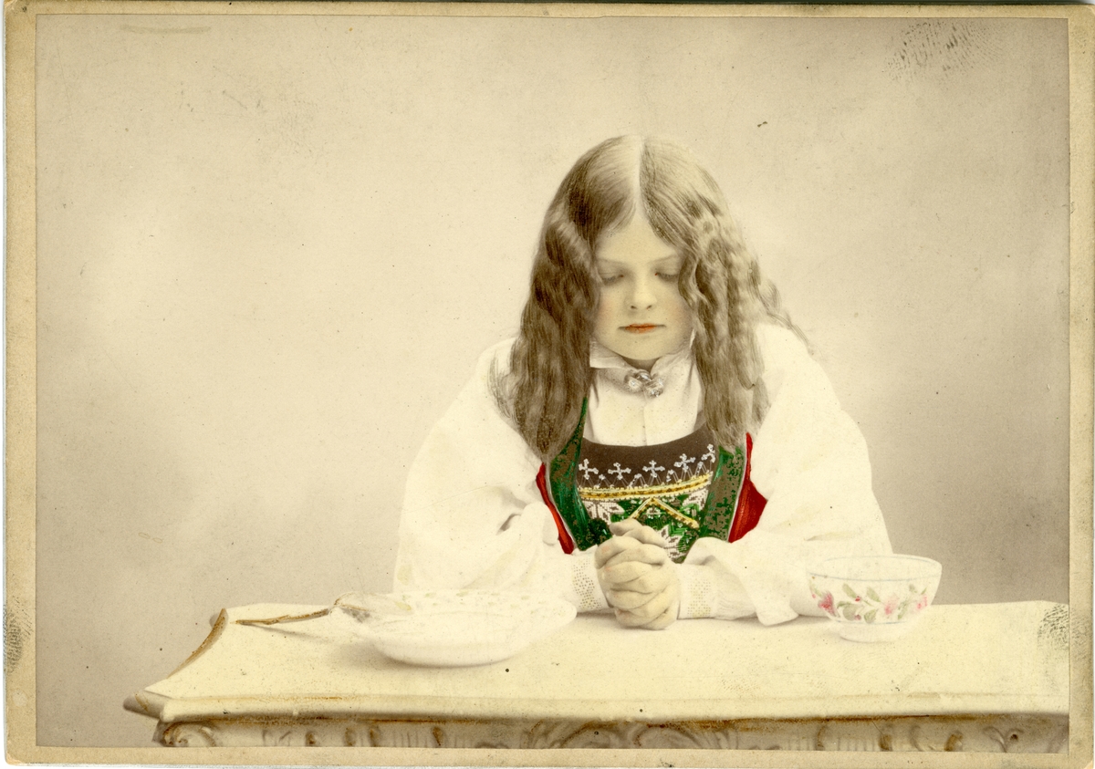 Kolorert studiofotografi av jente med Hardangerdrakt, sitter med foldelde hender på bord.