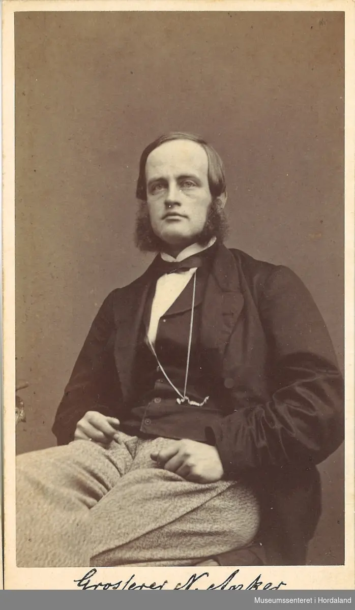 portrettfotografi av mann med "fipp"-skjegg, høgt hårfeste, svart jakke med vest og kvit skjorte, ljose bukser som sit på ein stol