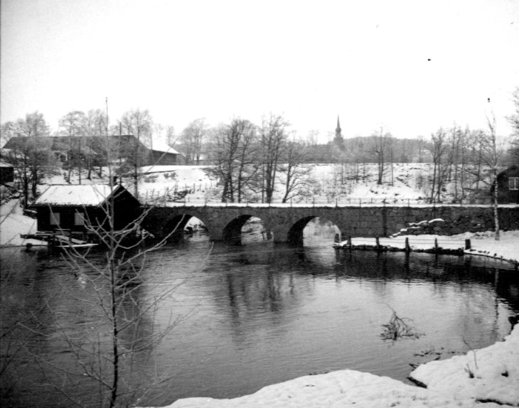 Bro över vattendrag. Bron är Vamme bro. Kyrktornet i bakgrunden tillhör Lerums kyrka.