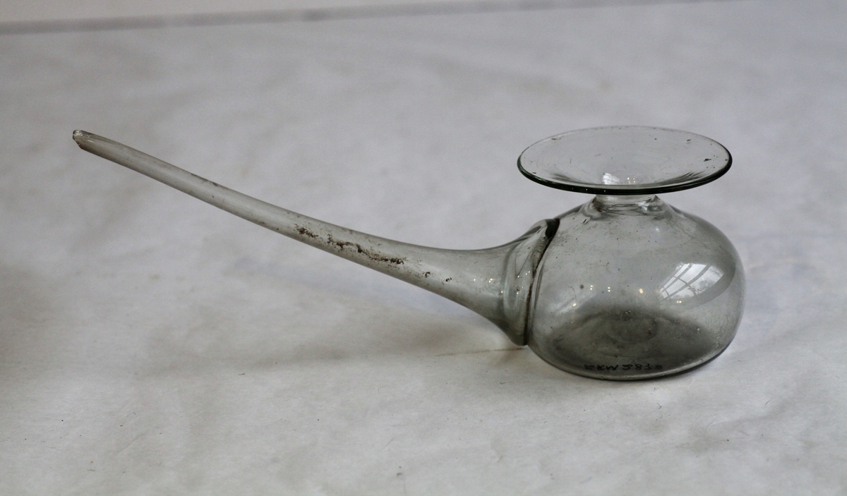 Brystglass med langt rør, brukt til pumping av morsmelk. Brystglass er laget av klar glassmasse. Tuten har sannsynlig vært  lenger.