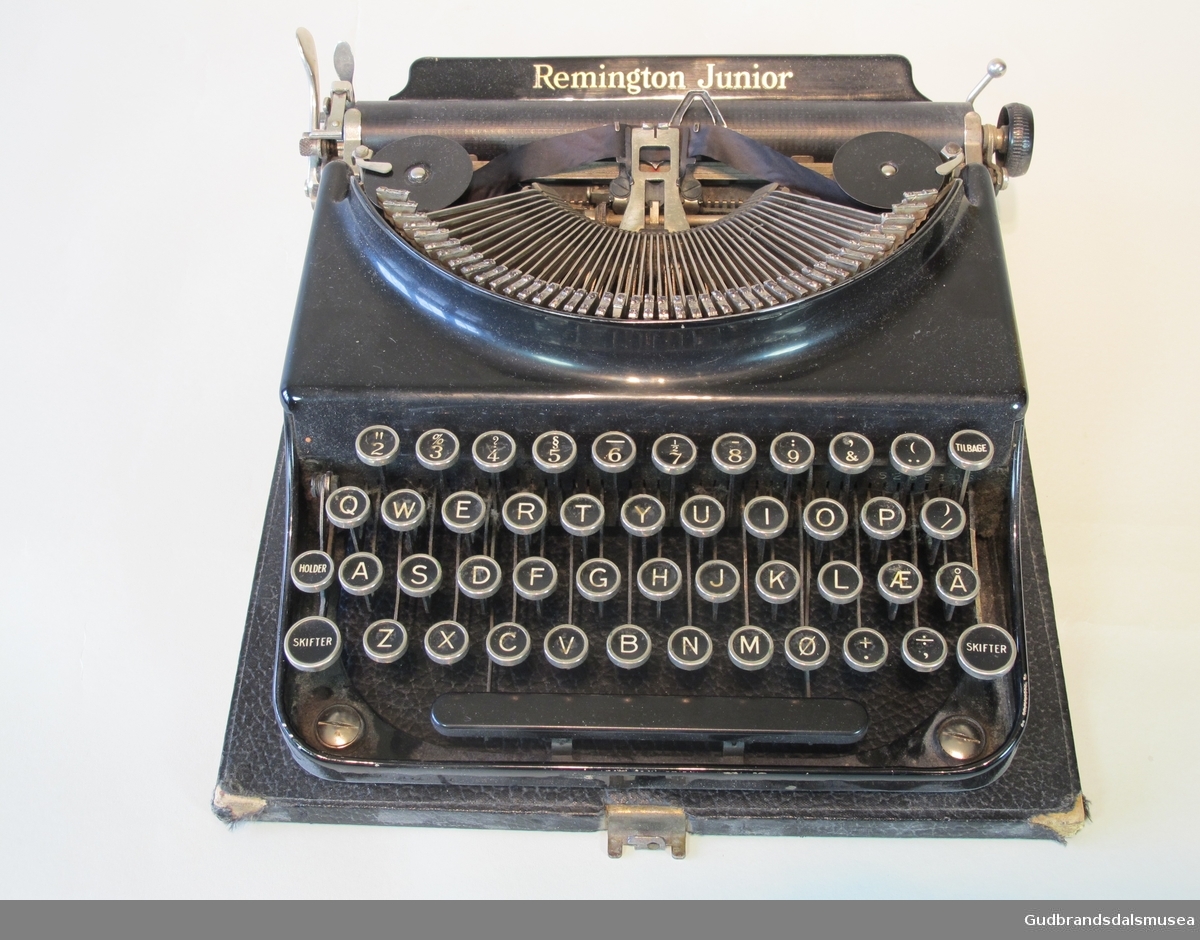 Manuell skrivemaskin med lokk. Har sort fargebånd. Norsk alfabet. Både maskin og lokk er noe slitt i kantene. Lokket har bærehåndtak.