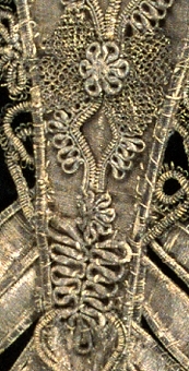 Bröstlapp i V-form, av siden med silvertråd broderat med läggsöm på bottenväven. 
Mönster: rankor och blommor lindade på band med silvertråd, flätade rosetter.
I ytterkanten lindade partier i olika uddmönster. Allt i mörknad silvertråd.
Baksida av gråvitt siden.
Anm: Har burits av Karl XII:s syster drottning Ulrika Eleonora d.y. 1688-1741