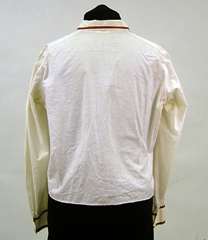 Blus sydd av tuskaftvävt bomullstyg. Stråveck sydda från axel på framstycket. Krage och manschetter är dekorerade med mönstervävt band, lika det band som är applicerat på förklädet.