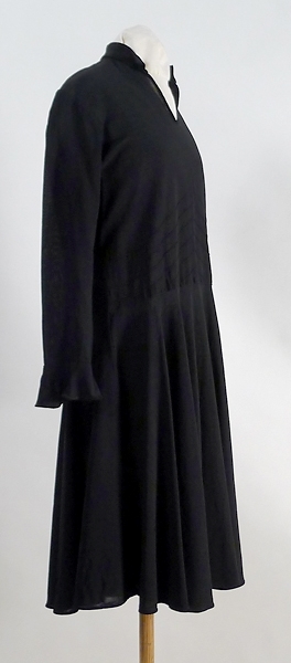 Flickklänning i  svart crepe av konstsilke med ståndkrage. Garneringsstickningar på framstycket samt bakstycket, helskuren kjol.