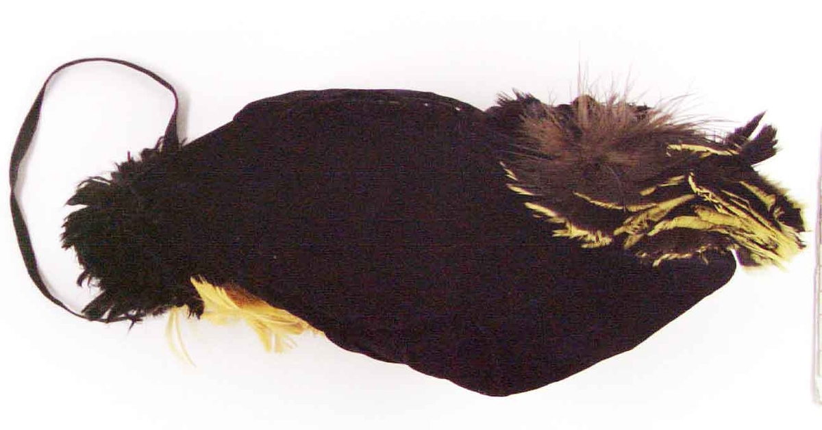 Mössa av svart sammet fodrad med gult bomullstyg. I sidorna svarta och gula fjädrar. Ett svart knytband. Hör till cirkusdräkten 30356.