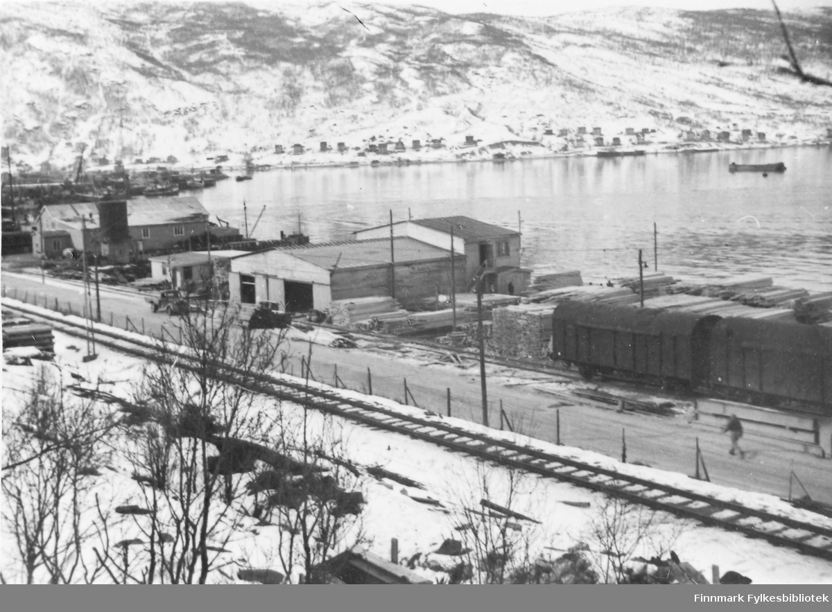 Før og etter 1944, Nordland, Troms. Oversiktsbilde over del av trelastanlegg. Vi ser et høvleri medtilhørende kontorer. Alt situert på kaiområdet. Jernbanen passerer iforgrunn. I bakgrunn av bildet på andre siden av sjøen kan vi se noeav bybildet i Narvik. Sneen ligger ennå.