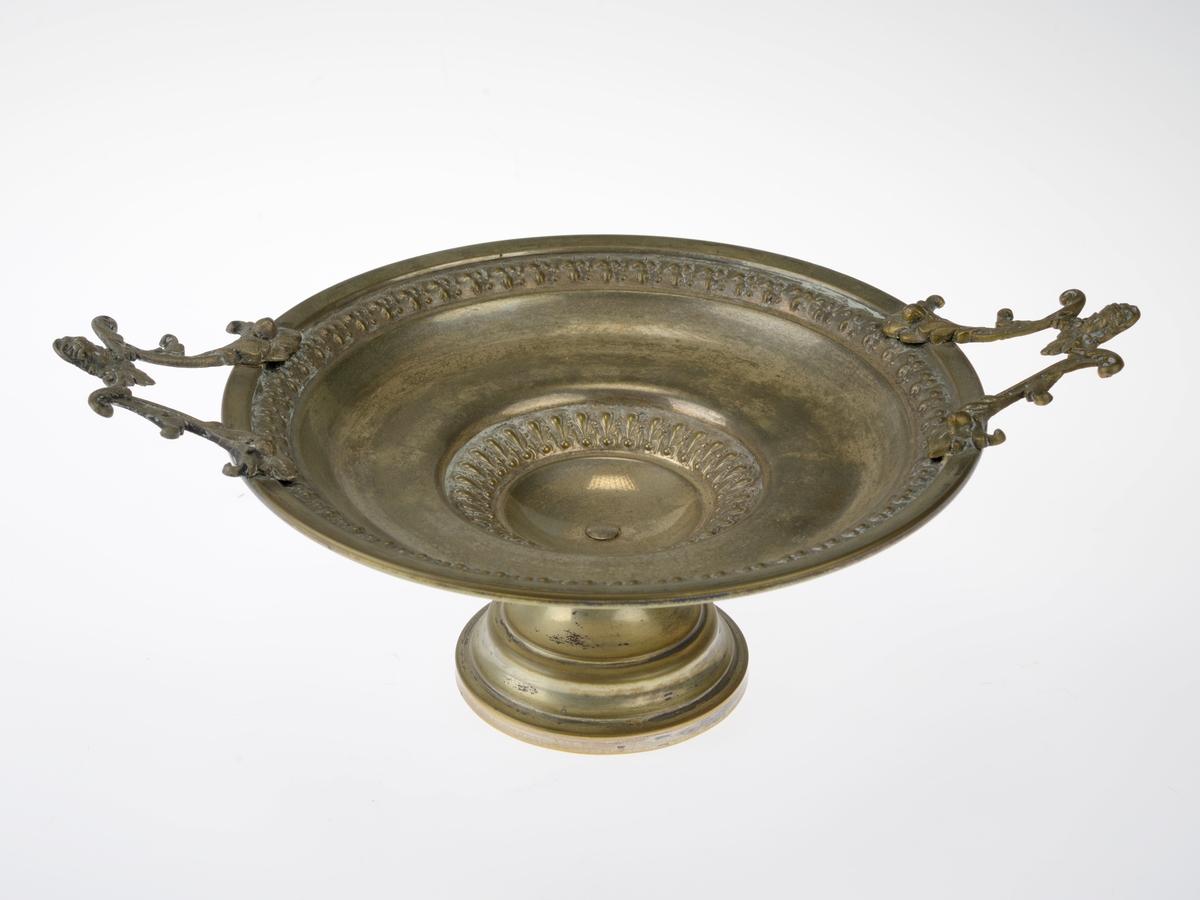 En skål (eller et fat) på stett. Skålen har utstanset mønster i bunnen og langs randen. Skålen er skrudd på en fot. På hver side er det skrudd på håndtak. Håndtakene er av et annet metall en skålen. Utformingen på håndtakene er to ranker som holdes sammen av et lite ansikt/hode.