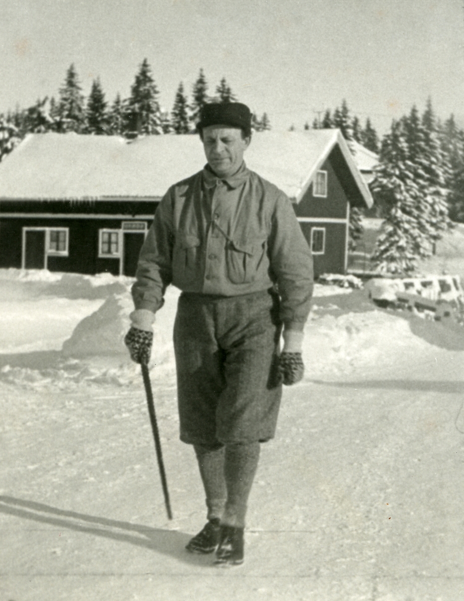 Haakon Garaasen (6/7 1887 - 1957) ute og spaserer med stav. Uthus el. lign. i bakgrunnen. Vinter.