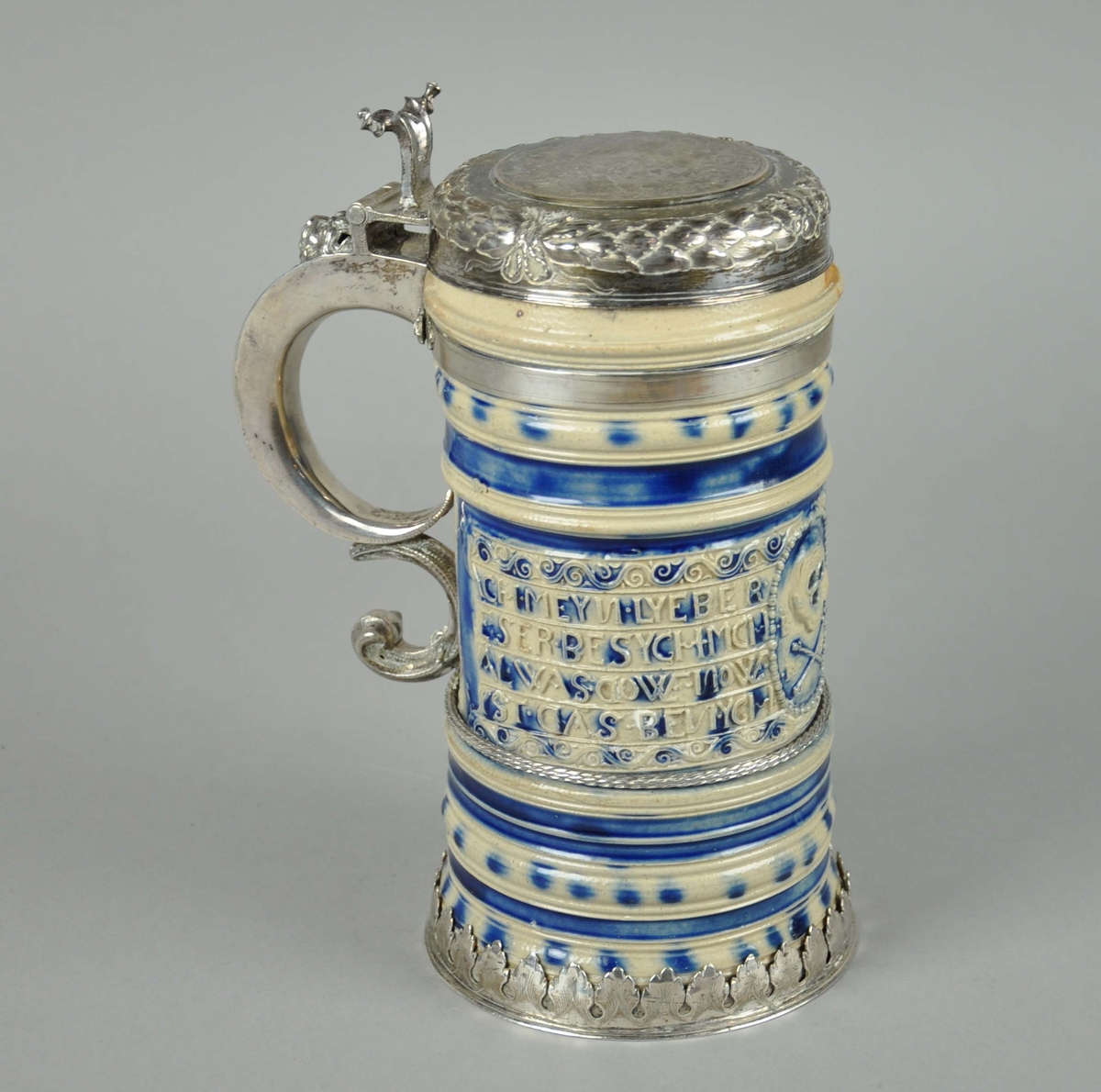 Stentøy ølkanne med sølvhank og sølvlokk. Hank og lokk er sekundære. På siden gjengitt et dødninghode med korslagte bein og en lengre innskrift på tysk. Sølvhank med dyremaske og sølvlokk med eikeløvskrans og gravert våpenskjold. Våpenskjoldet er familien Monrads våpen. Initiale FM og årstallet 1689 kan tyde på at det er Frederik Monrad, sogneprest i Øyer. Han var gift med Birgitte Cold. Initiale BCh kan være hennes, selv om navnet normalt ikke skrives med Ch.
Det tyder på at kannen opprinnelig har hatt hank av stentøy, som i 1689 er erstattet med sølvhank.