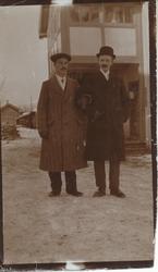 To unge menn utenfor bygning. Frakker, lue og bowlerhatt.