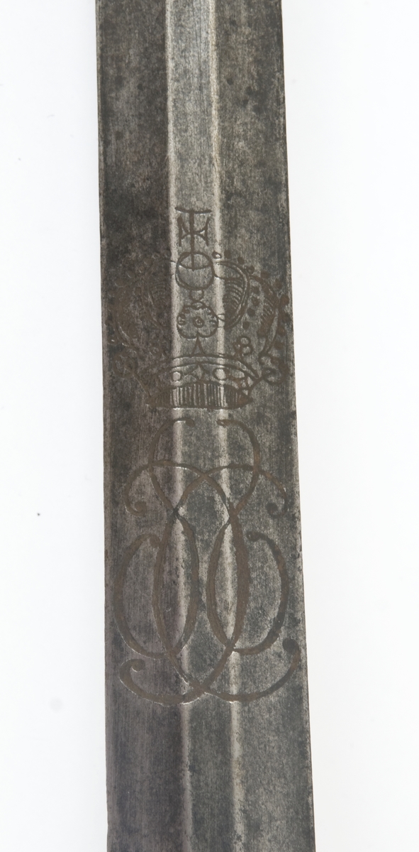 Offiserskorde dansk/norsk modell 1710. Messingfeste. Plommeformet knapp med nitteknapp. Hjerteformede parerplater, Fremre parerstang går over i håndbøylen som er tappet inn i knappen, rett bakre parerstang som ender i en flattrykt dråpe. Tvunnet messingtråd grep med messingbånd avslutning oppe og nede. Tveegget, sekskantet, rett klinge. Christian VI kronede speilmonogram på begeg sider av klingen.