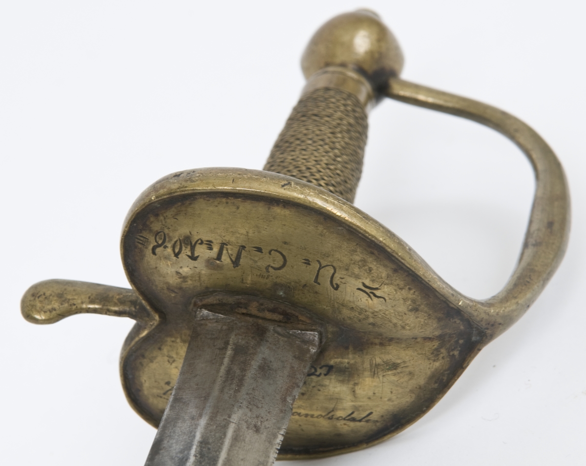 Offiserskorde dansk/norsk modell 1710. Messingfeste. Plommeformet knapp med nitteknapp. Hjerteformede parerplater, Fremre parerstang går over i håndbøylen som er tappet inn i knappen, rett bakre parerstang som ender i en flattrykt dråpe. Tvunnet messingtråd grep med messingbånd avslutning oppe og nede. Tveegget, sekskantet, rett klinge. Christian VI kronede speilmonogram på begeg sider av klingen.