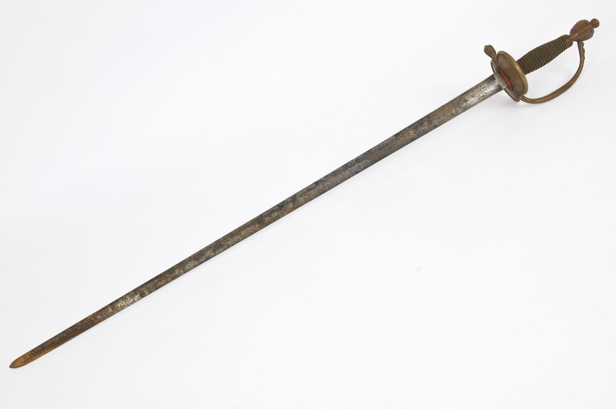 Dansk/Norsk underoffiserskårde modell 1777 for artilleriet. Messing feste. Vaseformet knapp med nitteknapp. Tregrep med snodd messingtråds bevikling og  messingring oppe og nede. Nyreformede parerplater. Rett bakre parerstang ender i en blomsterknopp. Fremre parerstang går over i håndbøylen. Nyreformede parerplater med kantlinje. 
Tveegget,  rett klinge med ovalt tverrsnitt på klingebrystet. Klingen har ryggstillt midtås.. Rester av blånering og forgyllt dekor  på begge sider. bl a det Dansk/Norske riksvåpen. Militære atributter og merket VIVAT øverst på klingebrystet.
