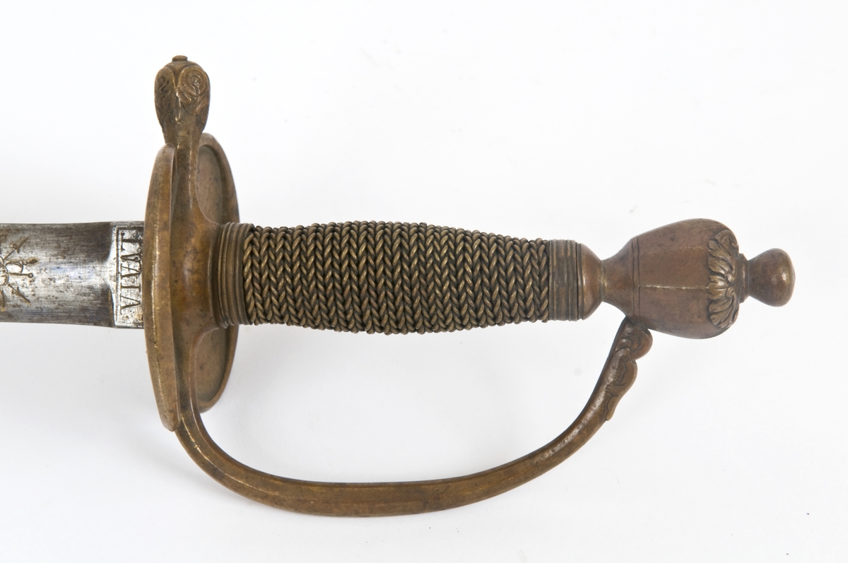 Dansk/Norsk underoffiserskårde modell 1777 for artilleriet. Messing feste. Vaseformet knapp med nitteknapp. Tregrep med snodd messingtråds bevikling og  messingring oppe og nede. Nyreformede parerplater. Rett bakre parerstang ender i en blomsterknopp. Fremre parerstang går over i håndbøylen. Nyreformede parerplater med kantlinje. 
Tveegget,  rett klinge med ovalt tverrsnitt på klingebrystet. Klingen har ryggstillt midtås.. Rester av blånering og forgyllt dekor  på begge sider. bl a det Dansk/Norske riksvåpen. Militære atributter og merket VIVAT øverst på klingebrystet.