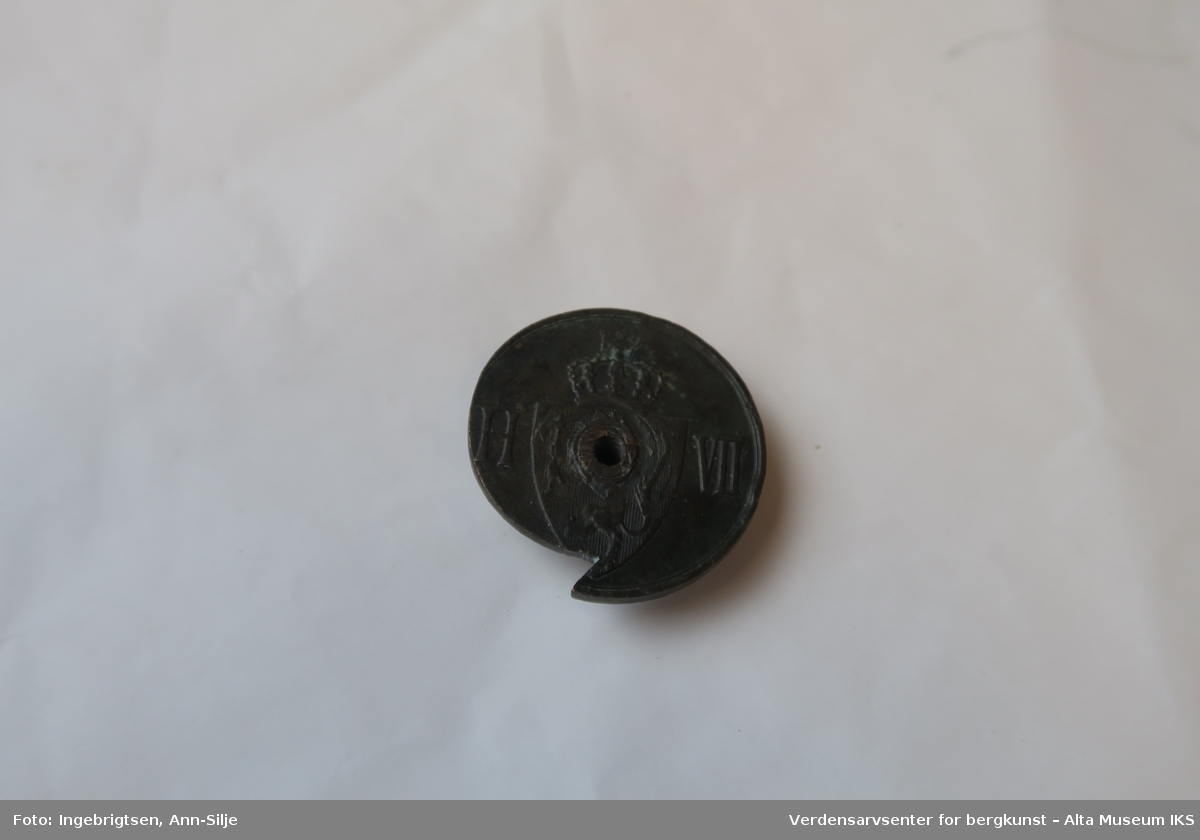 Form: Rund mynt med innskåret hakk og den er gjennomhullet. På den ene siden er det relieff med motiv av det norske riksvåpen med løve.