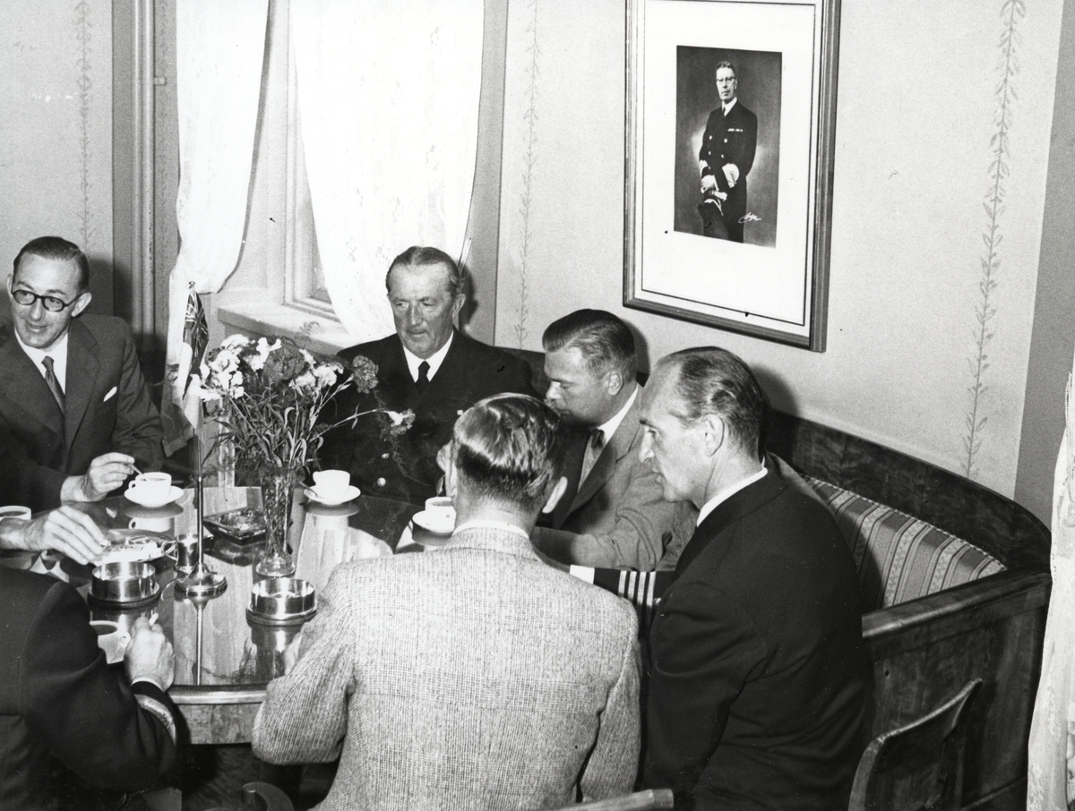 En grupp militärer sitter runt ett kaffebord. Vissa av dem är klädda i flottans uniformer, andra är kostymklädda. I mitten av bordet står en vas med blommor samt Engelska kungliga flottans flagga. En av männen är konteramiral Samuelson, en annan kommendörskapten av första graden Lind af Hageby.