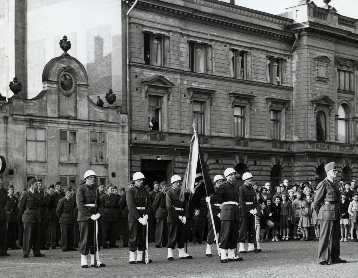 Uniformsklädda militärer från KA:2 står uppställda på Stortorget framför Karlskrona stadshotell. I bakgrunden framför hotellet står en folksamling bestående av barn och vuxna som tittar på.