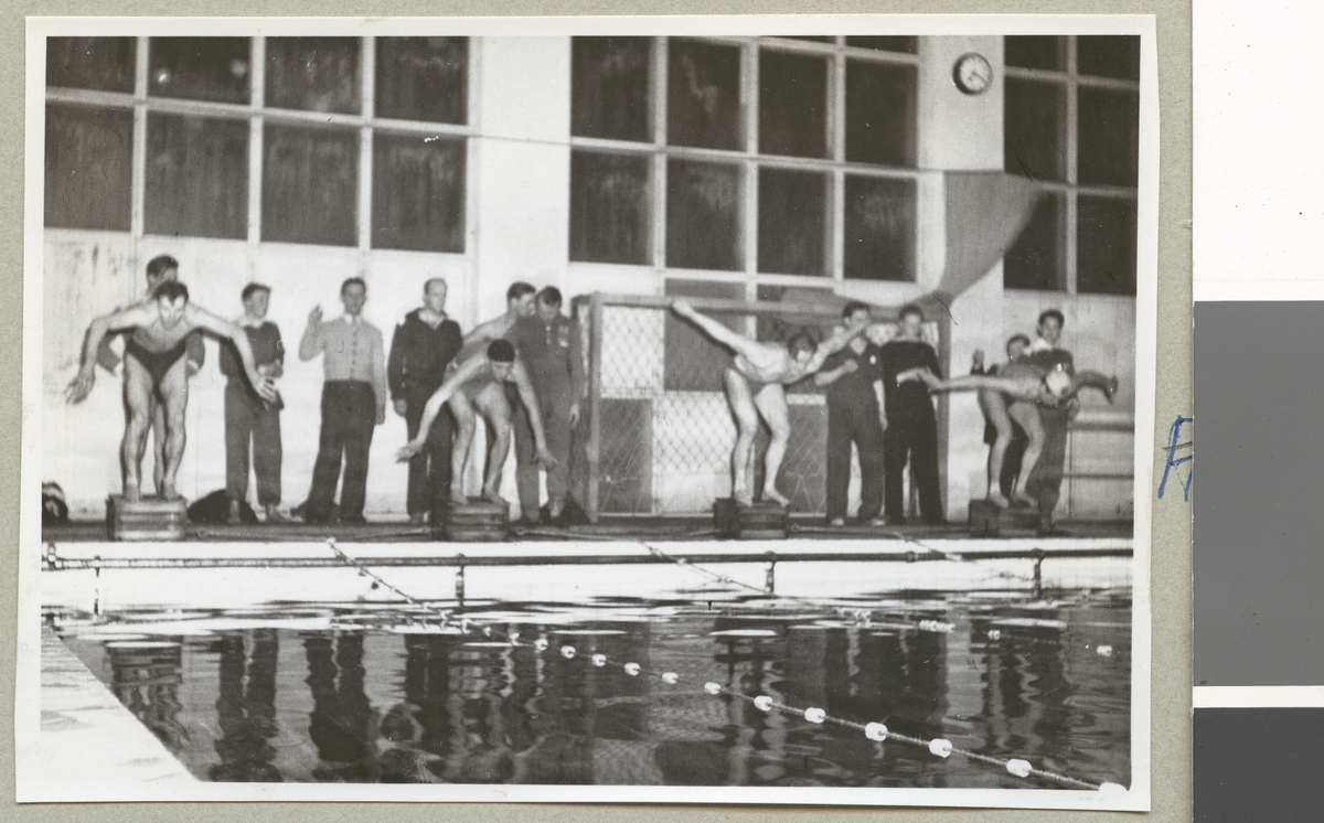 Fyra män i badkläder i färd med att hoppa i en simbassäng under en simtävling. Bakom dem står flera män och tittar på.