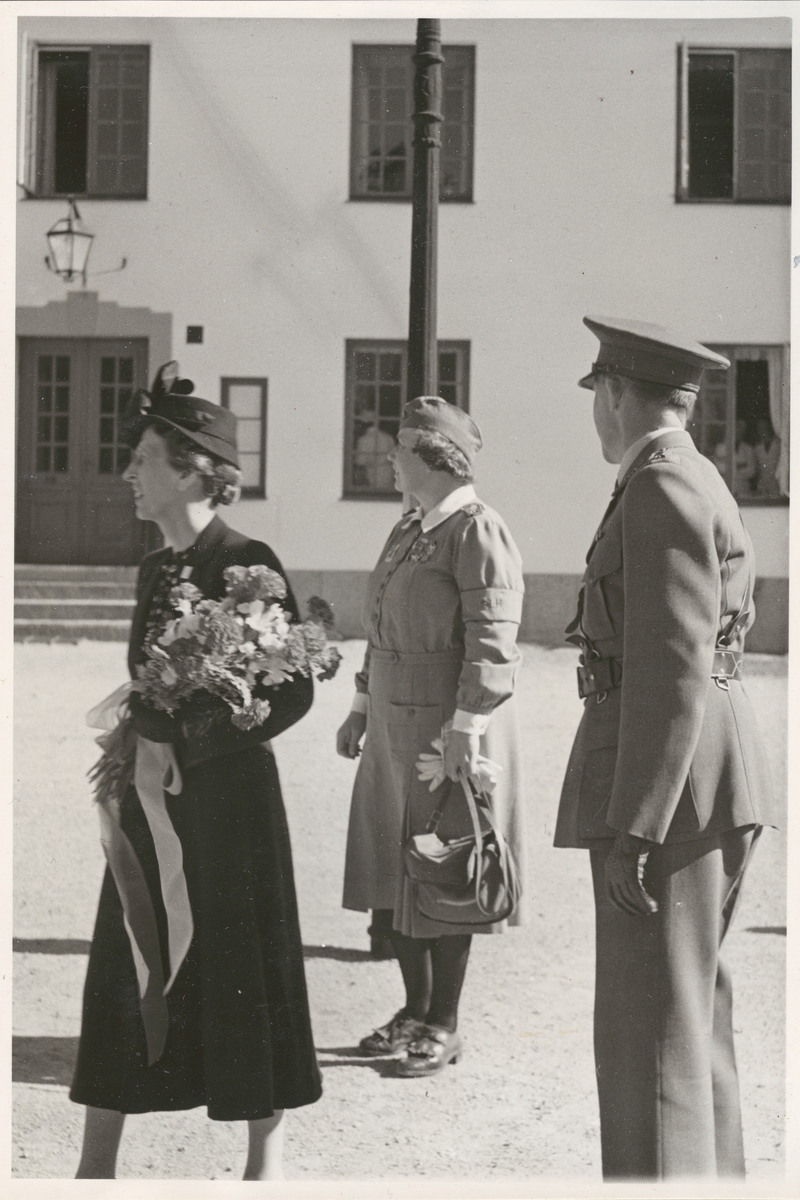 Drottning Louise, klädd i mörk dräkt och hatt samt med en blombukett i famnen, står utomhus på en grusyta tillsammans med en lotta och en militär från armén, båda i uniform. I bakgrunden syns det norra befälshuset på Kungsholmen med pusad fasad och småspröjsade fönster.