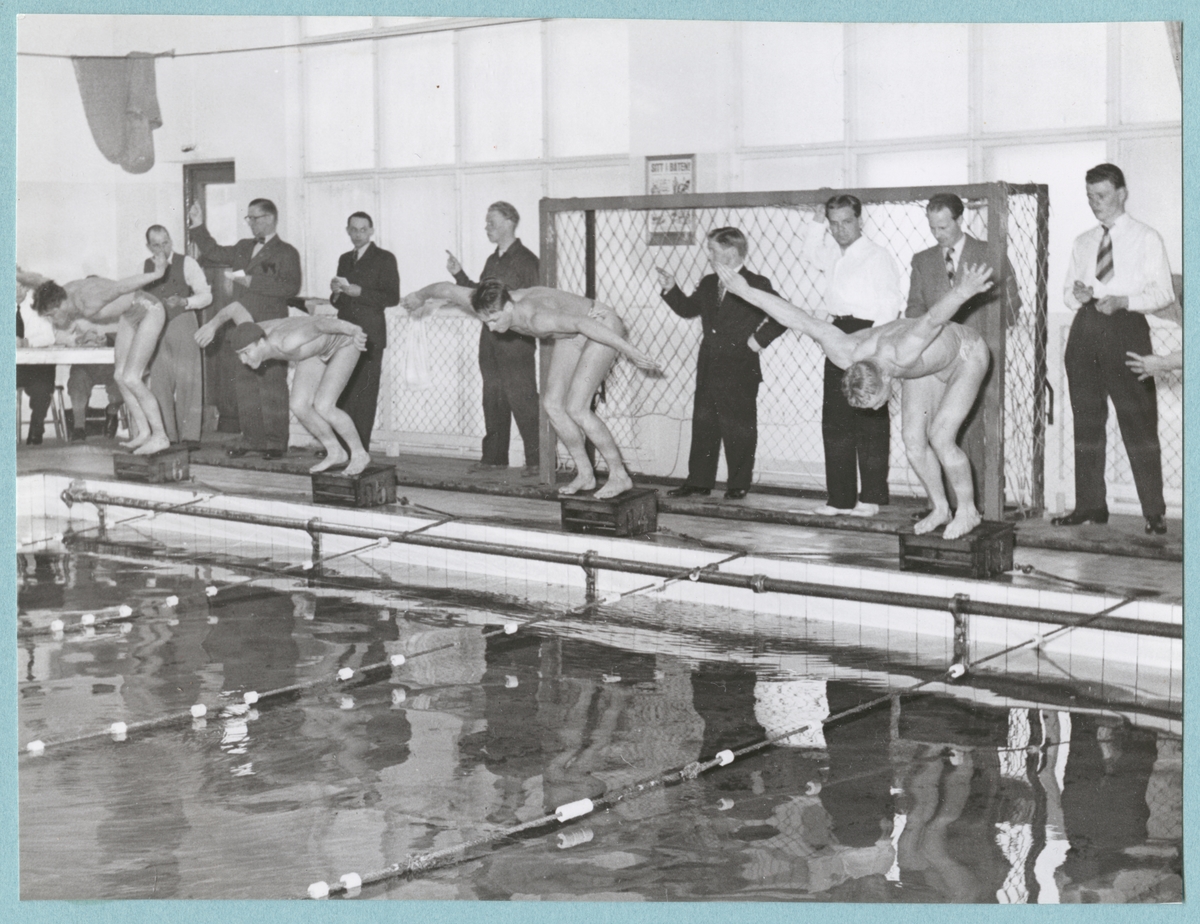 Fyra manliga simmare i badbyxor står på pallar och lutar sig fram för att hoppa i simbassängen. Bakom simmarna står ytterligare män klädda i skjorta alternativt kavaj. Intill väggen står ett mål. Fotot är daterat till 15-3-1952.
