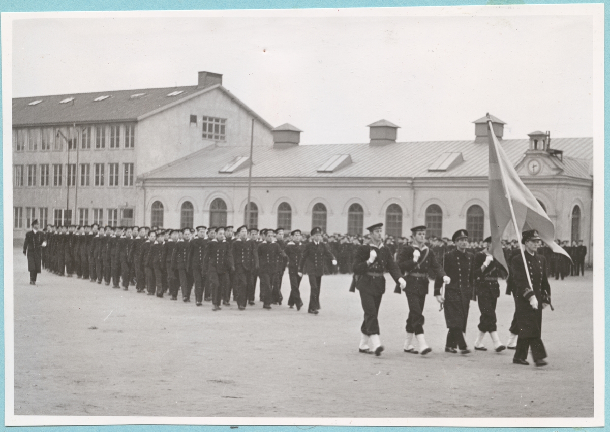 Militärmarsch med flottister på bataljon Sparres kaserngård. Framför gruppen går fyra högre flottister samt en fänrik bärandes på en svensk flagga. I bakgrunden syns Sparres excercishus.