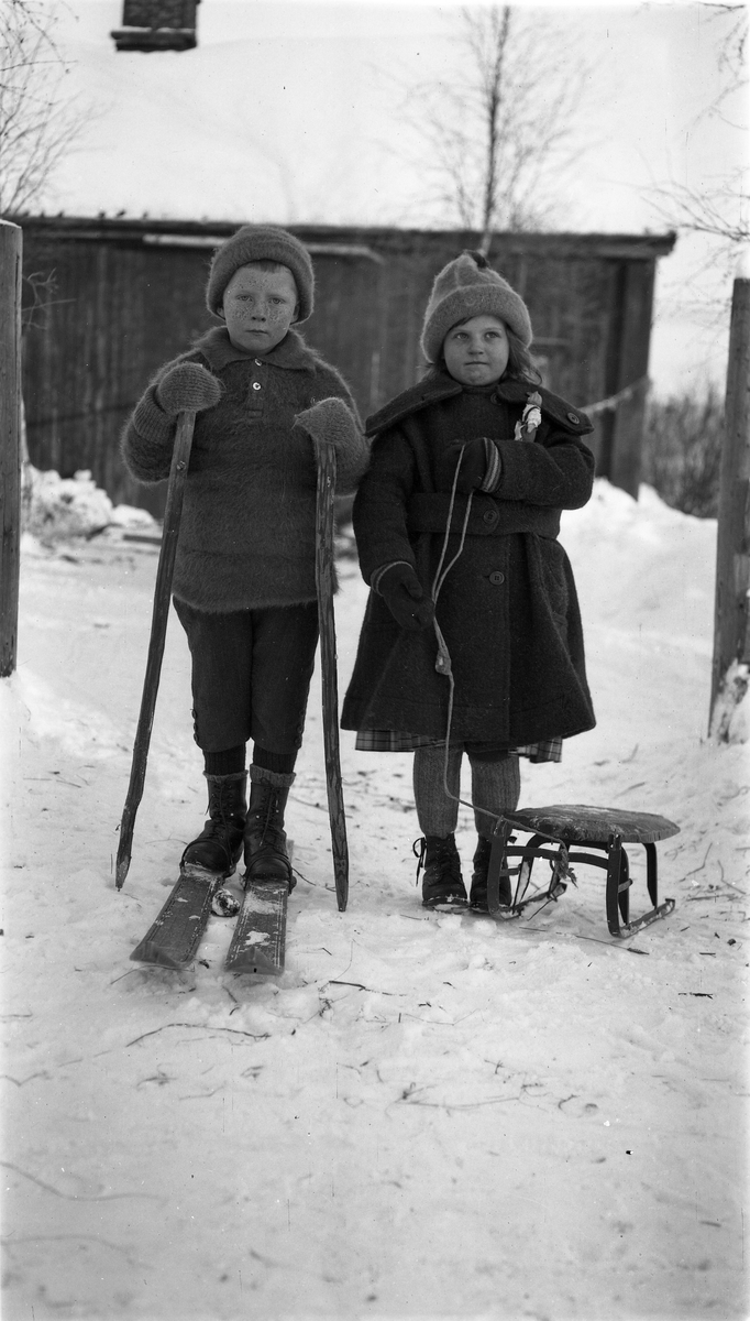 Vinterlek. To barn i helfigur, en gutt på ski, og ei jente med kjelke. Guttens navn er muligens Ole Moen.